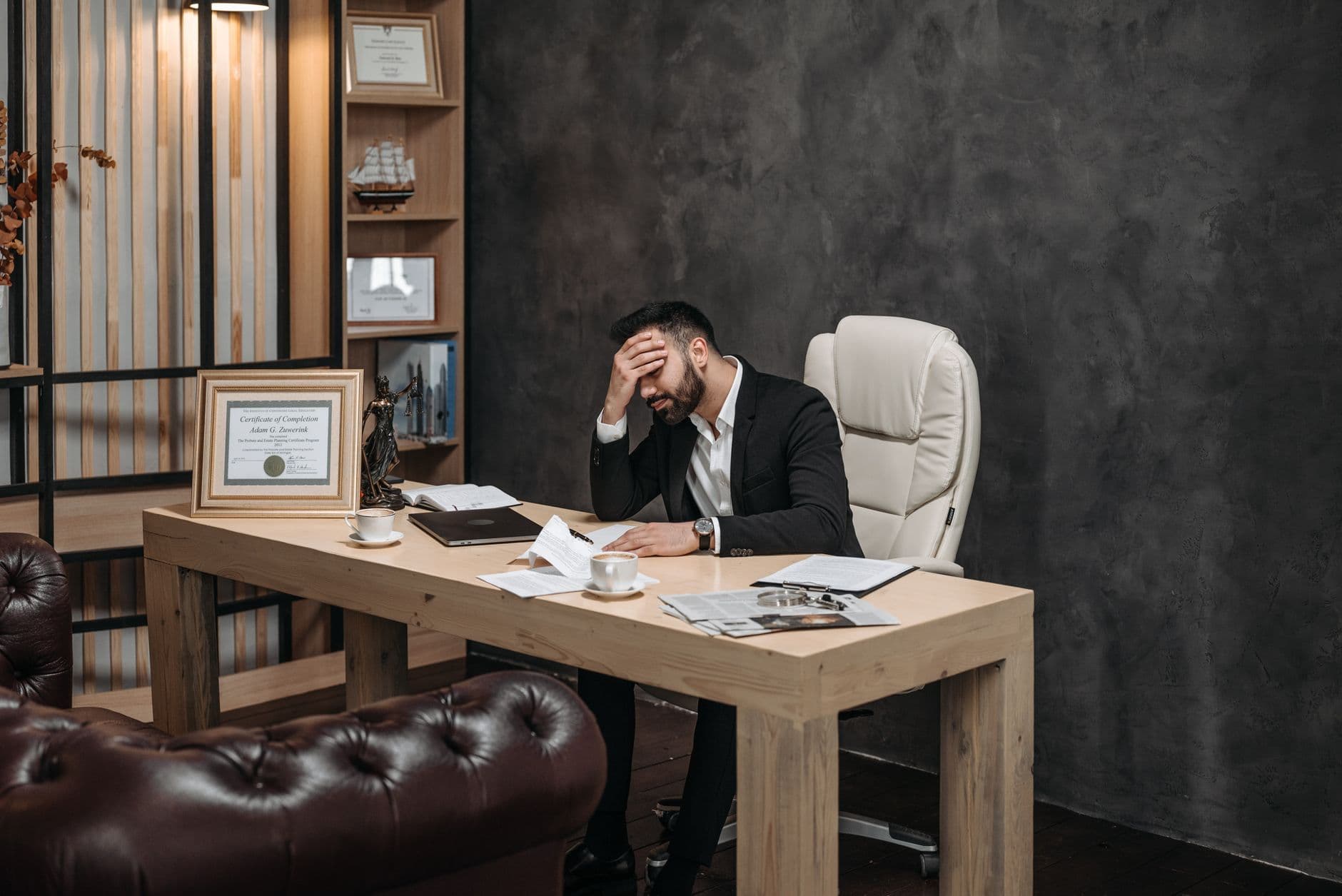Businessman in a black suit looking stressed at an office desk, holding his head with papers scattered around.