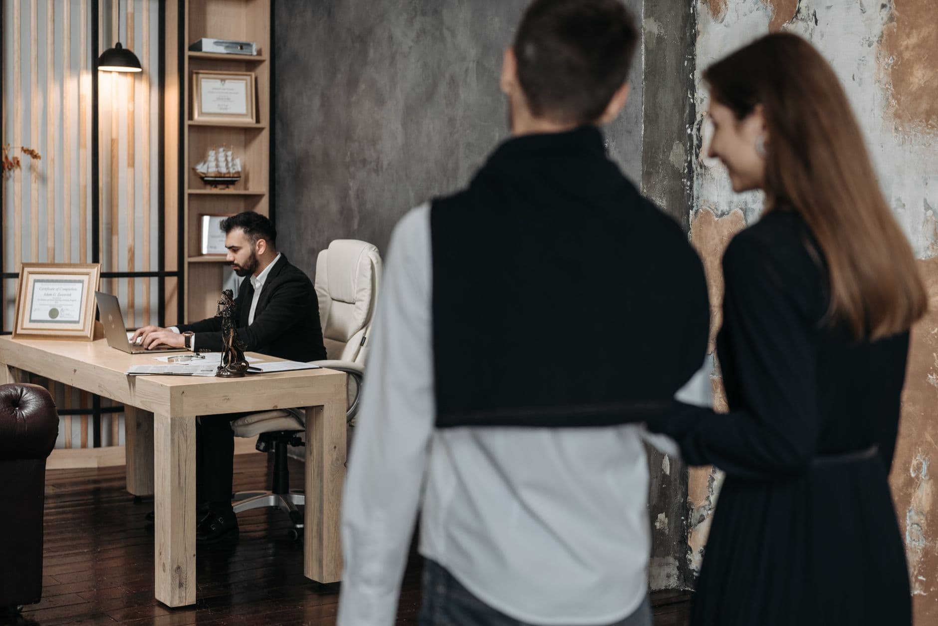 A business consultant working at a desk while a couple in formal wear approaches him in a modern office setting.