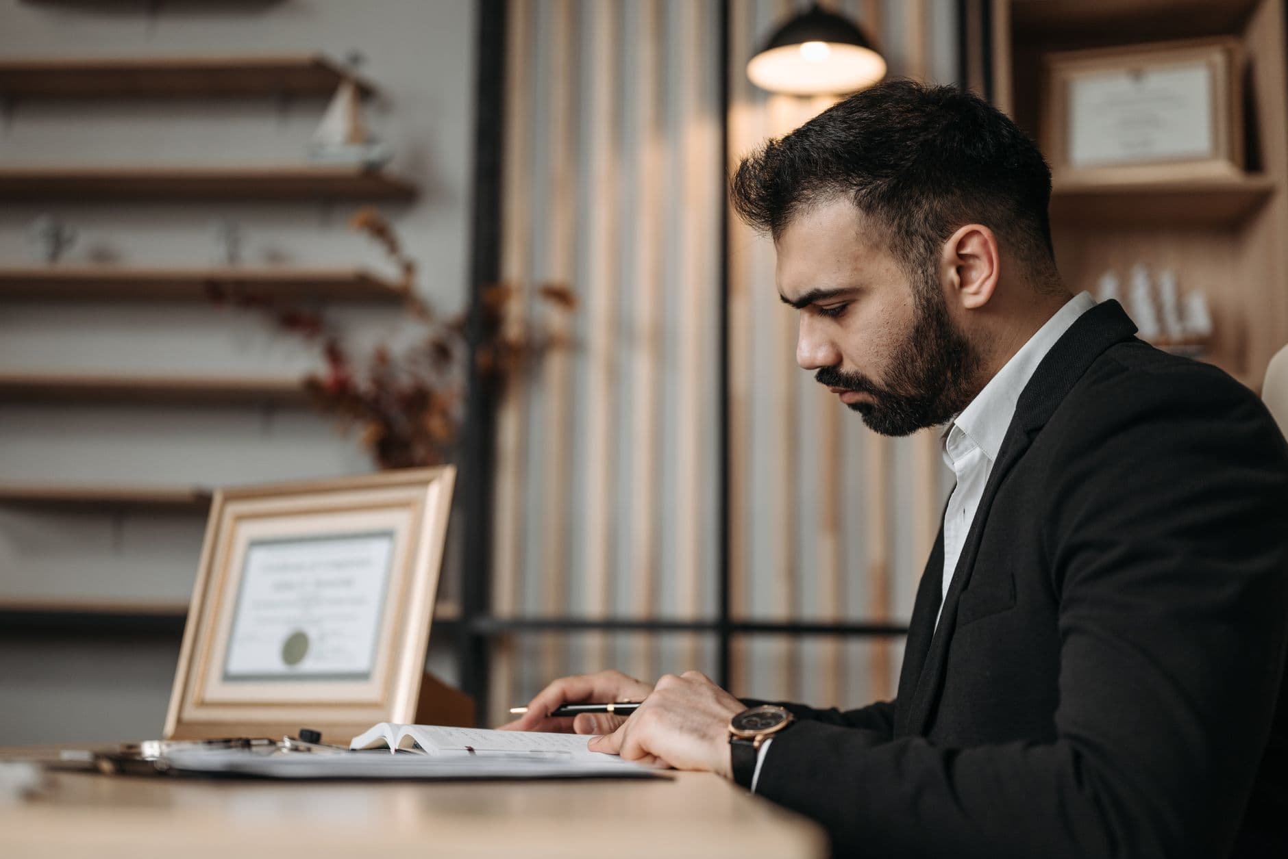 A professionally dressed man focused on writing at a desk within an office setting, suggesting a serious work environment.