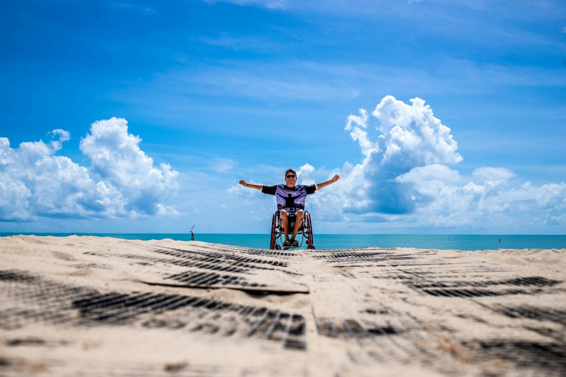 Man in wheelchair celebrating on João Pessoa beach with open arms under a clear blue sky.